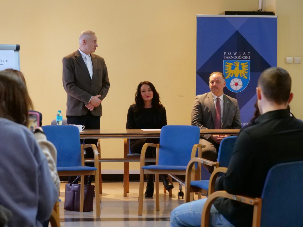 na pierwszym planie mężczyzna, stoi, przed nim stół, ubrany w koszule i marynarkę, obok siedzi kobieta, elegancko ubrana na czarno, obok kobiety mężczyzna w garniturze i krawacie, za nimi roll-up z herbem powiatu, przed nimi puste krzesła i fragment osób siedzących na widowni