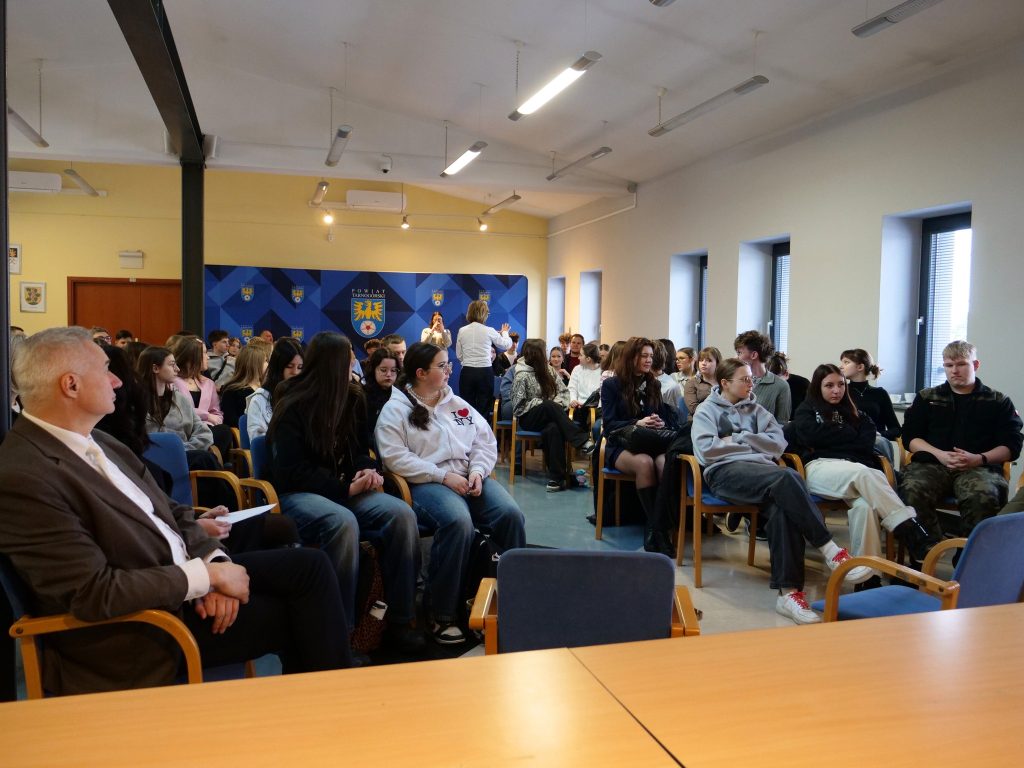 duża grupa uczniów w sali konferencyjnej, siedzą na krzesłach w rzędach, na widowni również kilku opiekunów, w tle widoczna ścianka z herbem powiatu
