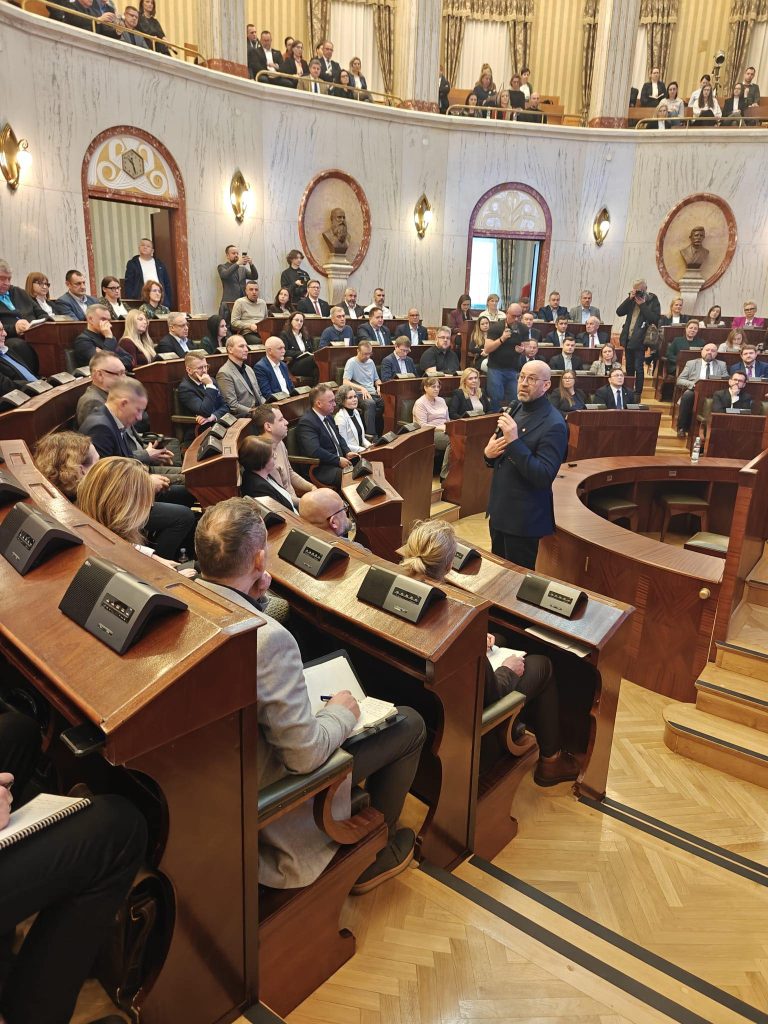 sala posiedzeń sejmowych, duża ilość osób siedzi na widowni, po prawej mężczyzna w garniturze stoi z mikrofonem w ręku, w tle ściany i rzeźby