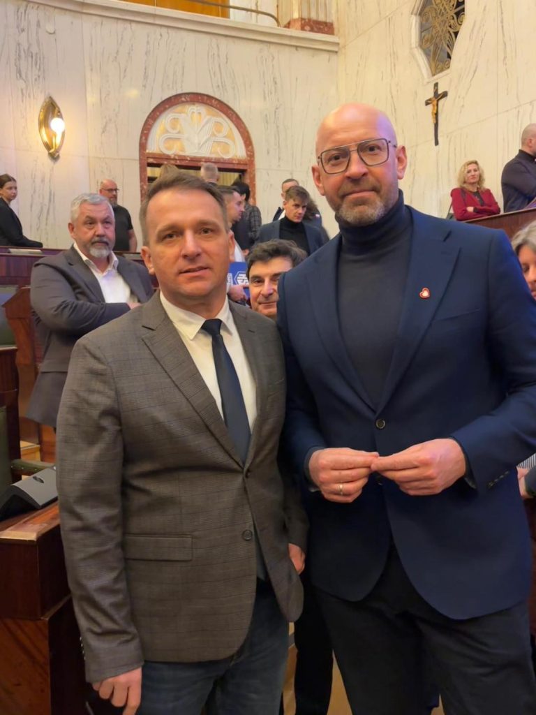 na pierwszy planie dwóch mężczyzn, elegancko ubranych w garnitury, w tle sala posiedzeń sejmowych