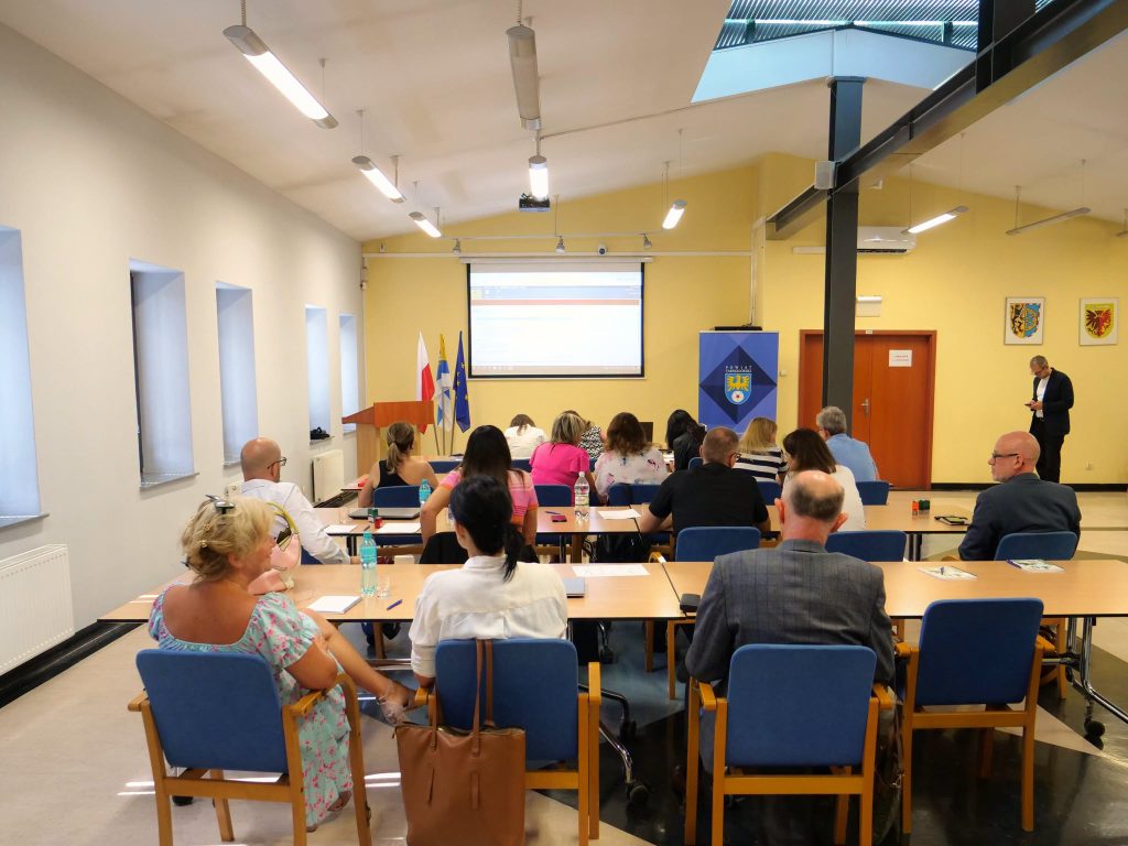 sala, na pierwszym planie osoby, siedzą na krzesłach, tyłem, przy stołach, na dalszym planie kolejne rzędy stołów i krzeseł oraz siedzących osób, po prawej ściana z okiennicami, w tle ściana, na ścianie wisi ekran, po prawej drzwi